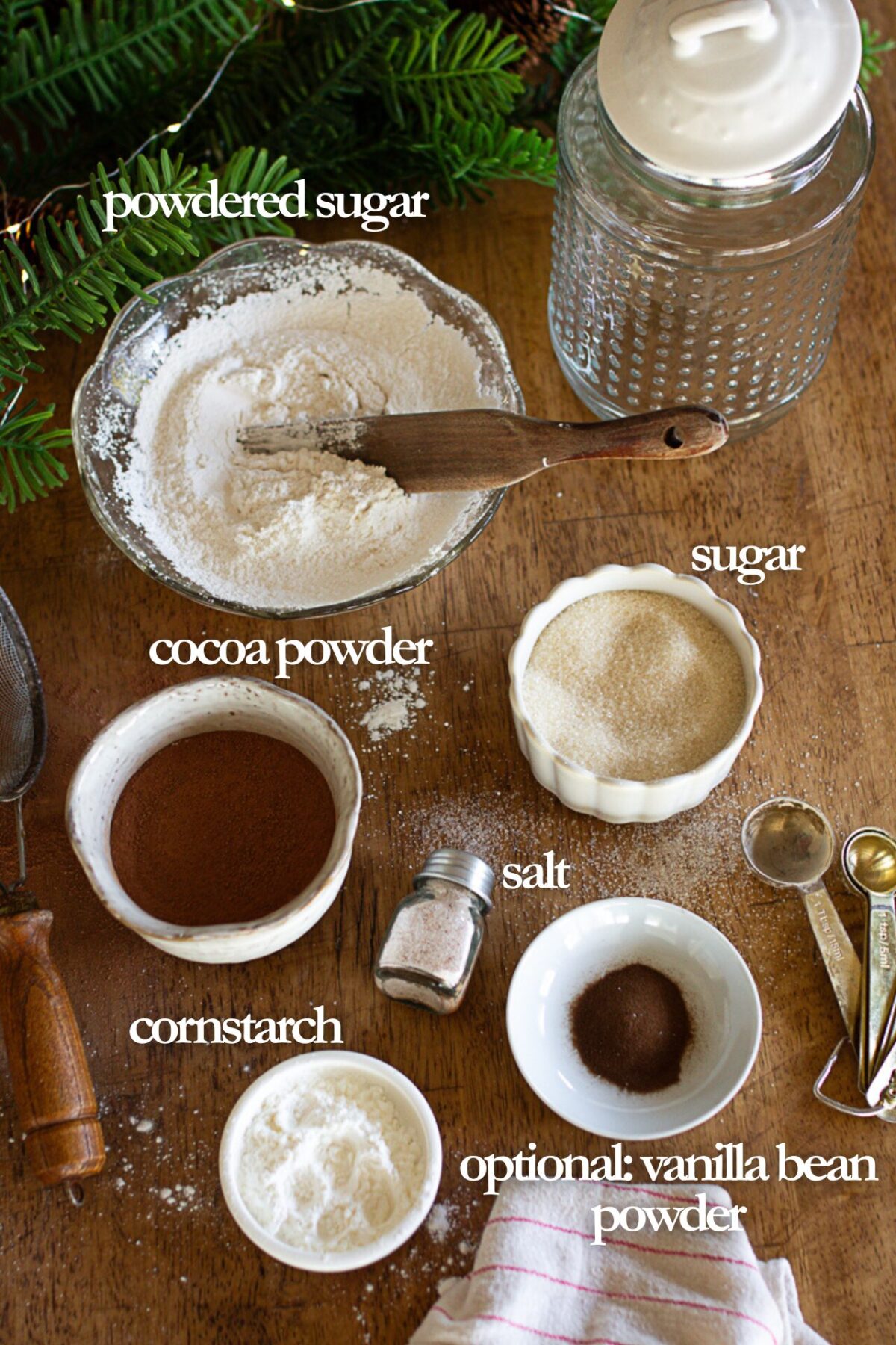 ingredients list for hot cocoa: powdered sugar, regular sugar, cornstarch, salt, cocoa power, and optional vanilla bean powder, all spread out on a table.