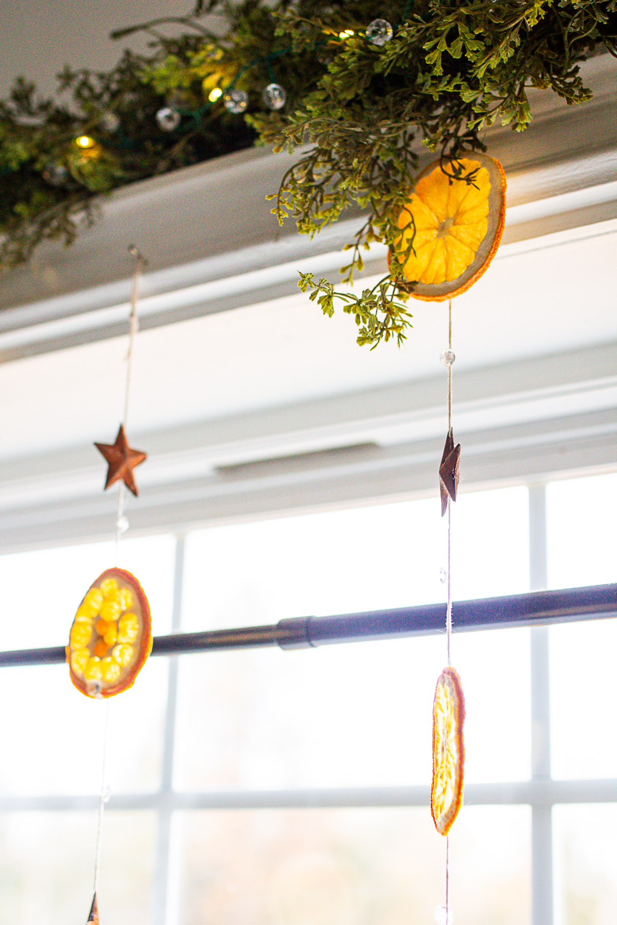 dried oranges hanging from strings above a window thats covered with faux cedar garland.