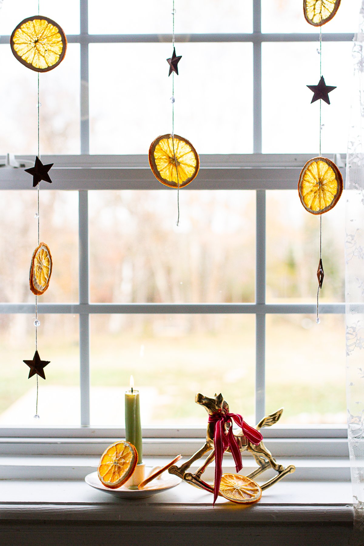 Orange and tin star garlands handing from a window above a brass rocking horse figurine and candle.