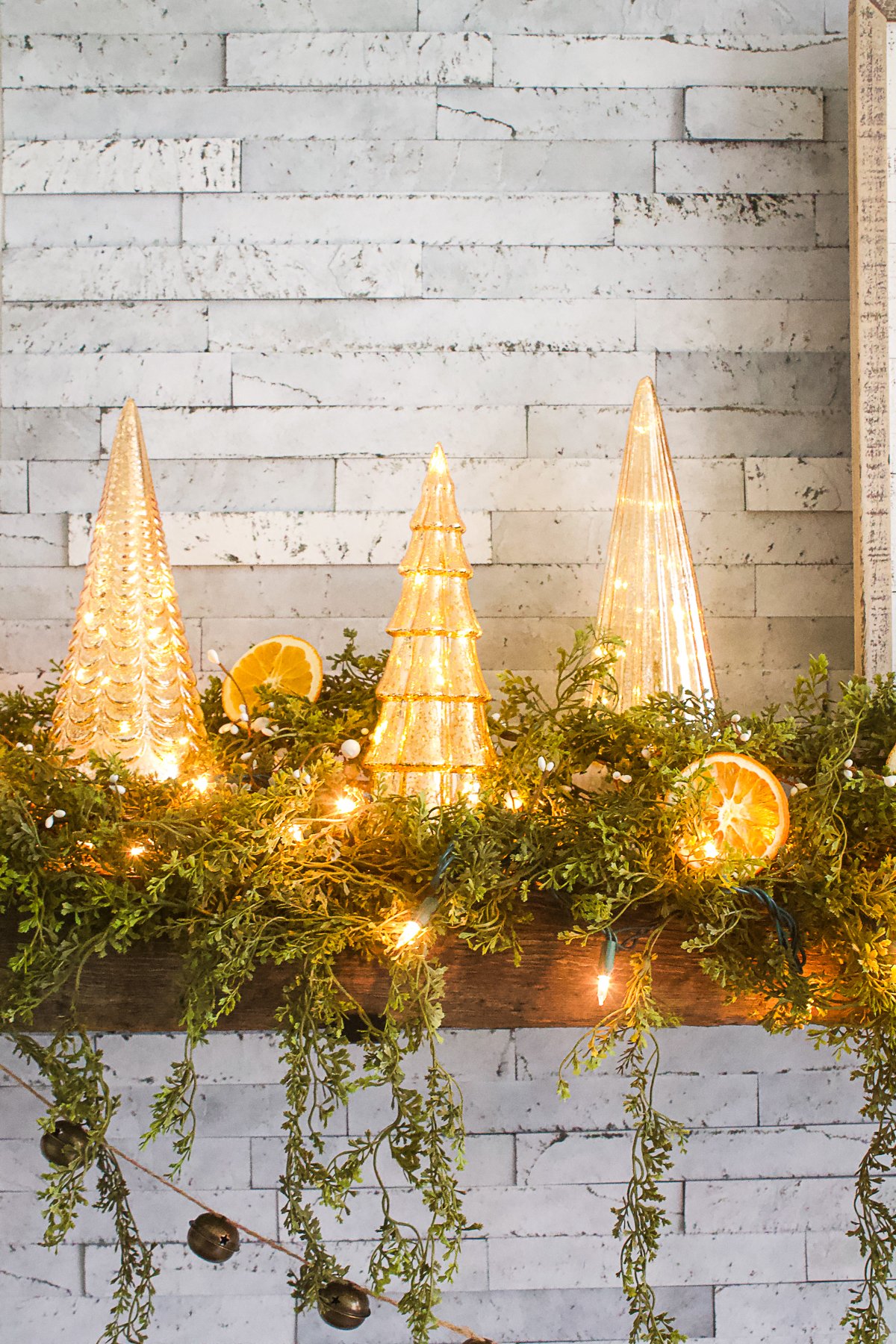 Three Mercury glass trees on mantle surrounded by faux cedar greens and dried oranges. 