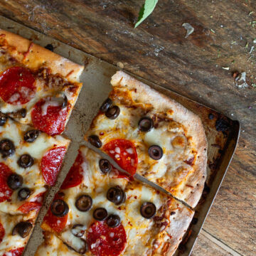 A wide angled, close-up shot of a baked homemade sheet pan pepperoni and black olive pizza, cut into slices, fresh from the oven, styled with fresh sage and whole garlic bulbs on a dark wood background