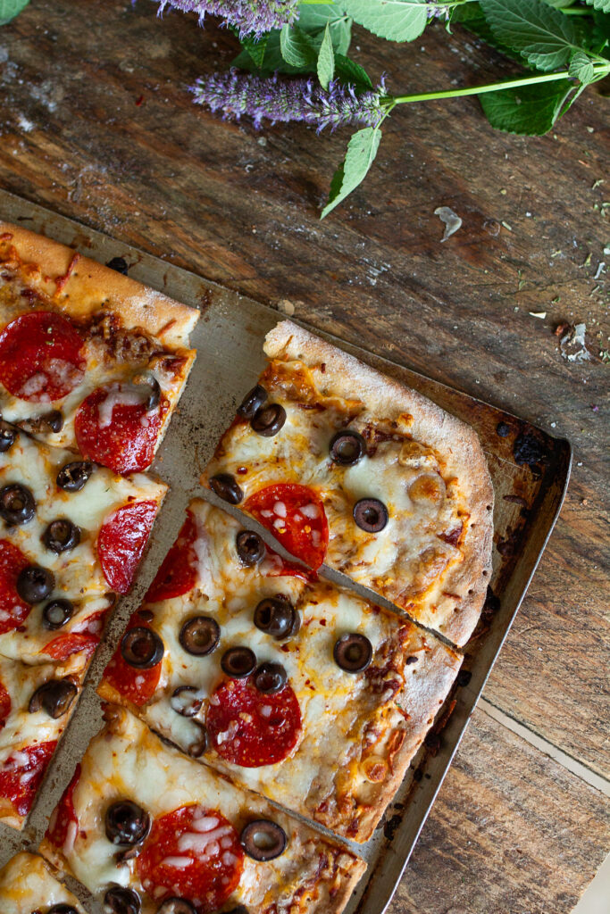 A wide angled, close-up shot of a baked homemade sheet pan pepperoni and black olive pizza, cut into slices, fresh from the oven, styled with fresh sage and whole garlic bulbs on a dark wood background