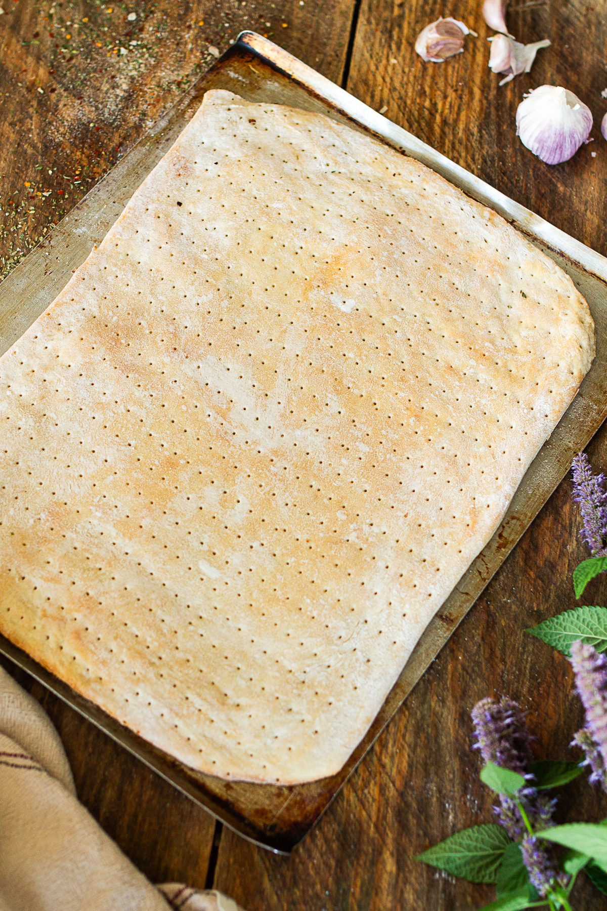 A rustic, rectangular cooked pizza dough docked with a rolling docker tool on an old baking sheet, styled on a dark wood table with garlic bulbs and purple flowers.