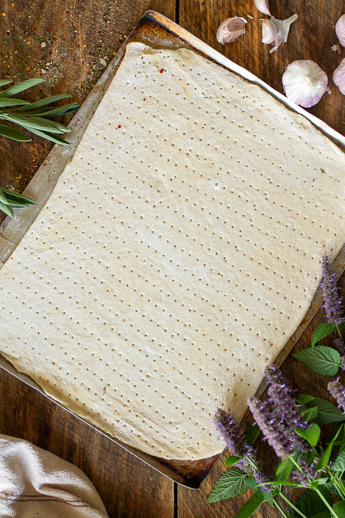 A rustic, rectangular raw pizza dough docked with a rolling docker tool on an old baking sheet, styled on a dark wood table with garlic bulbs and purple flowers.