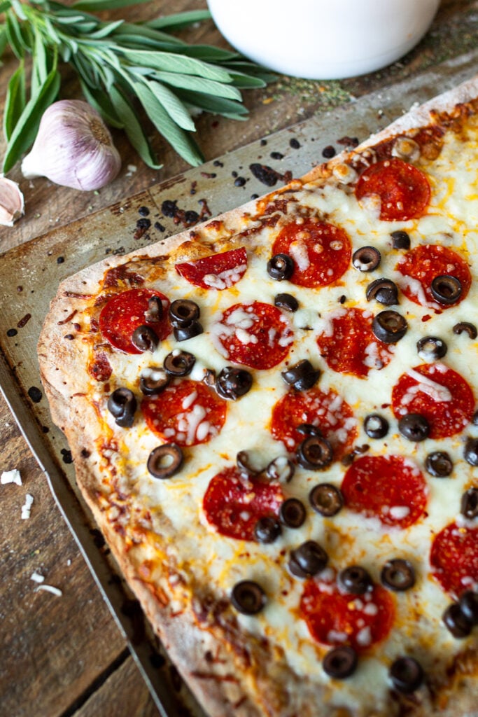 A wide angled, close-up shot of a baked homemade sheet pan pepperoni and black olive pizza fresh from the oven, styled with fresh sage and whole garlic bulbs on a dark wood background