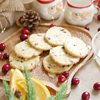 Cranberry orange cookies on a wooden ornament shaped cutting board with cranberries scattered around, Santa cocoa cups, and other Christmas decor.