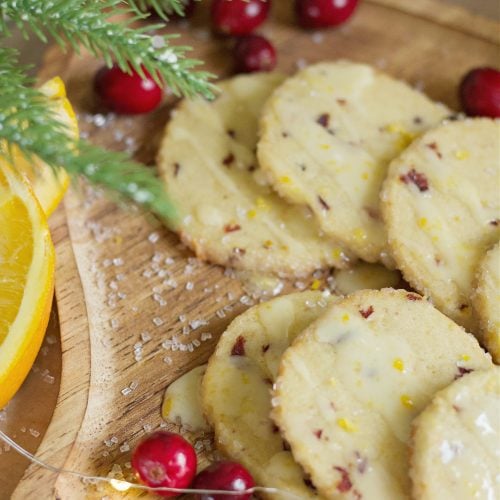 close up photo of cranberry orange icebox cookies on a wooden plate with oranges and cranberries scattered around them and twinkle lights.