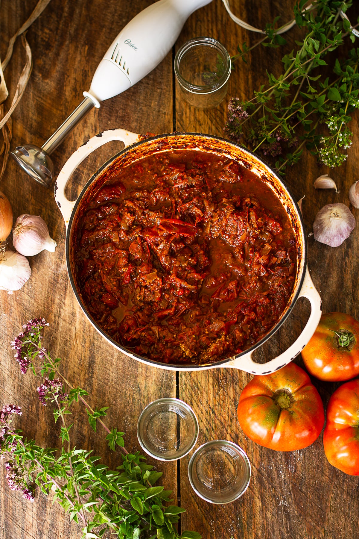 A large white enameled stockpot filled with chunky, slow-simmered homemade tomato sauce sitting on a wood surface next to an immersion stick blender and fresh herbs.