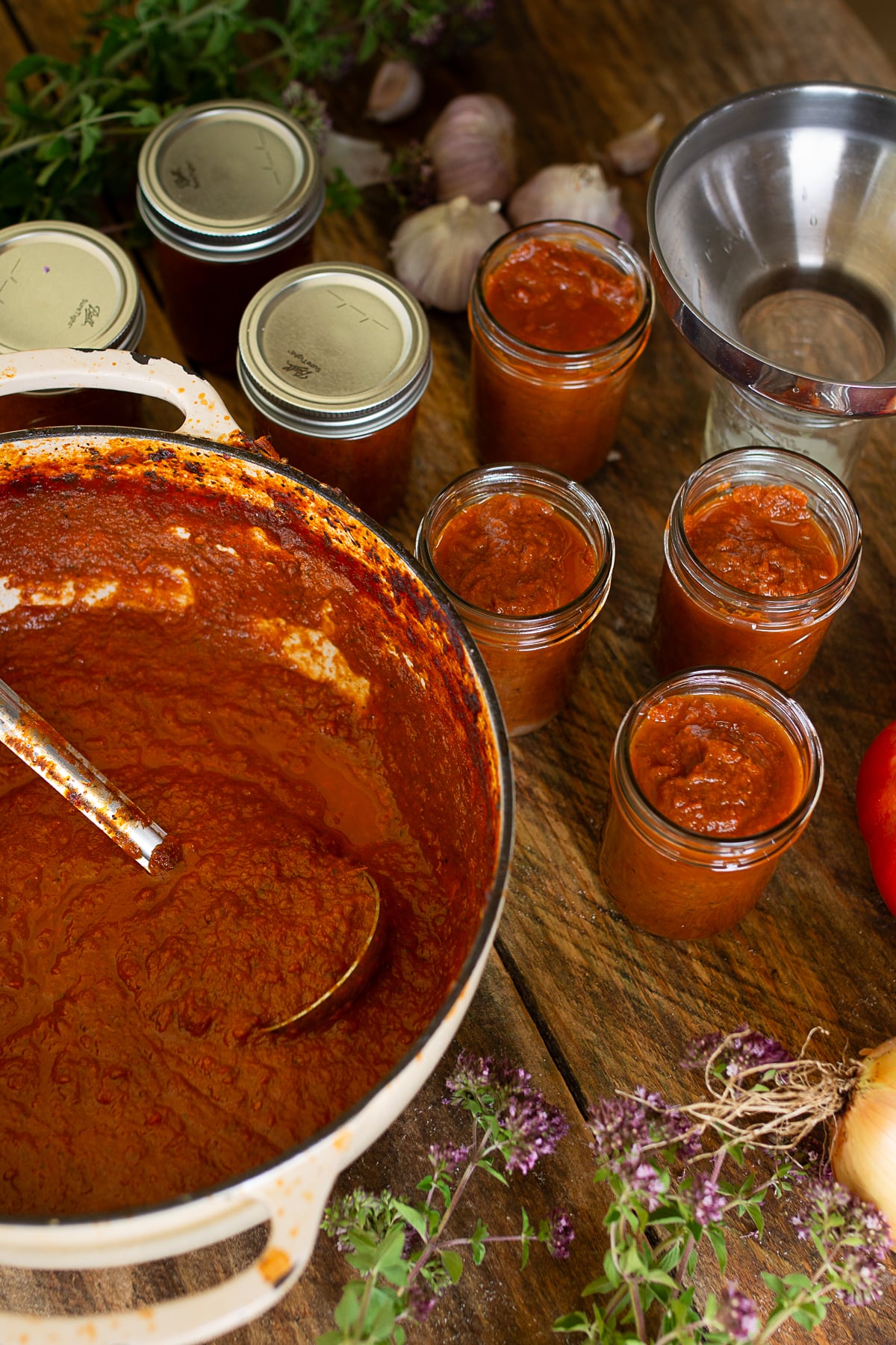 Ladling thick, hot homemade pizza sauce from a large pot into half-pint mason jars using a canning funnel, with sealed glass jars in the background.