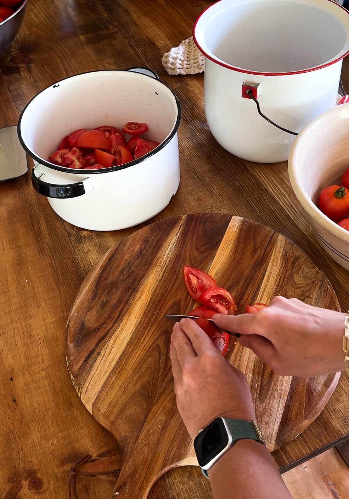 Hands cutting tomatoes with pot of cut tomatoes on the side. 