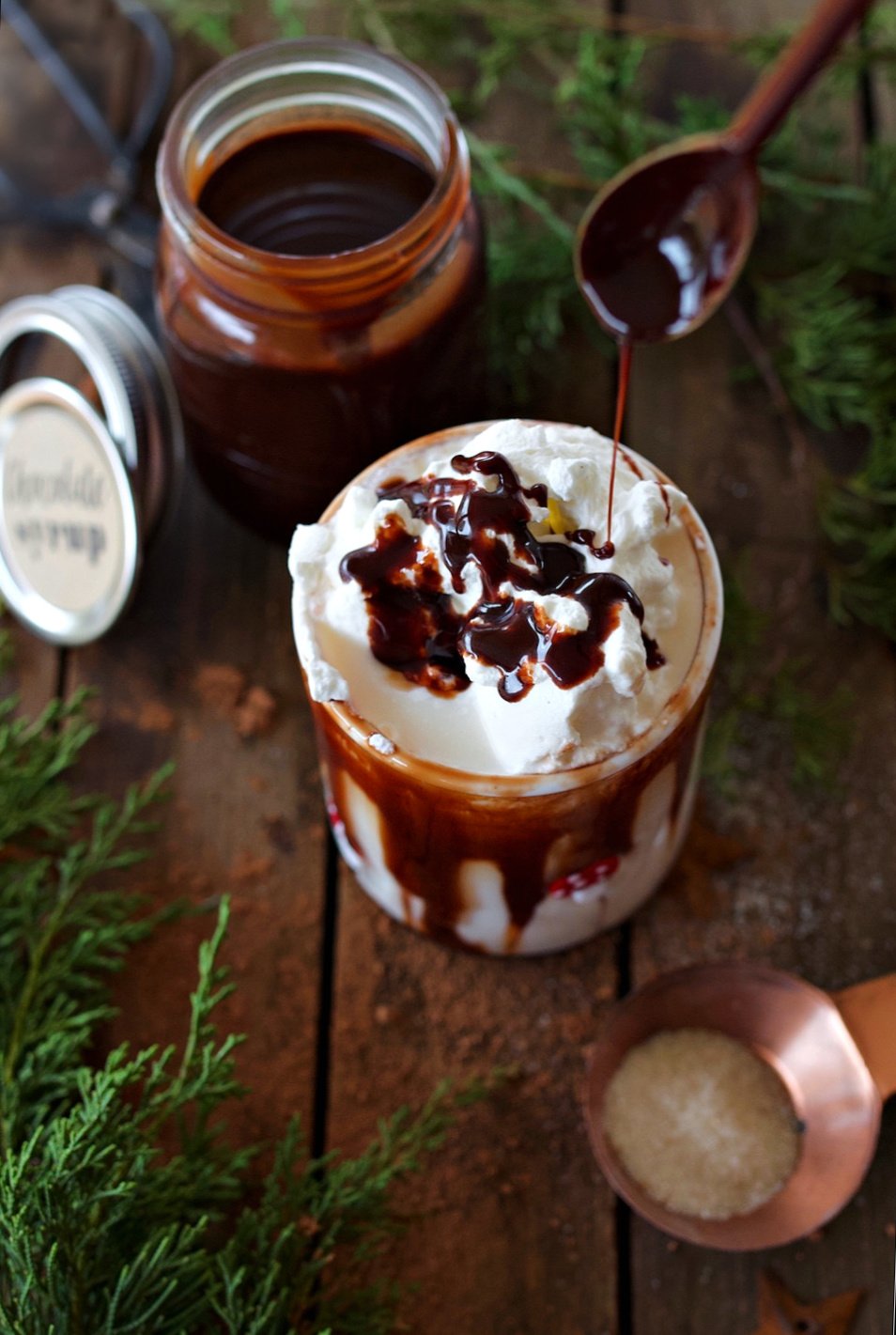 Cup of chocolate milk on counter with whipped cream on top being drizzled with homemade chocolate syrup.