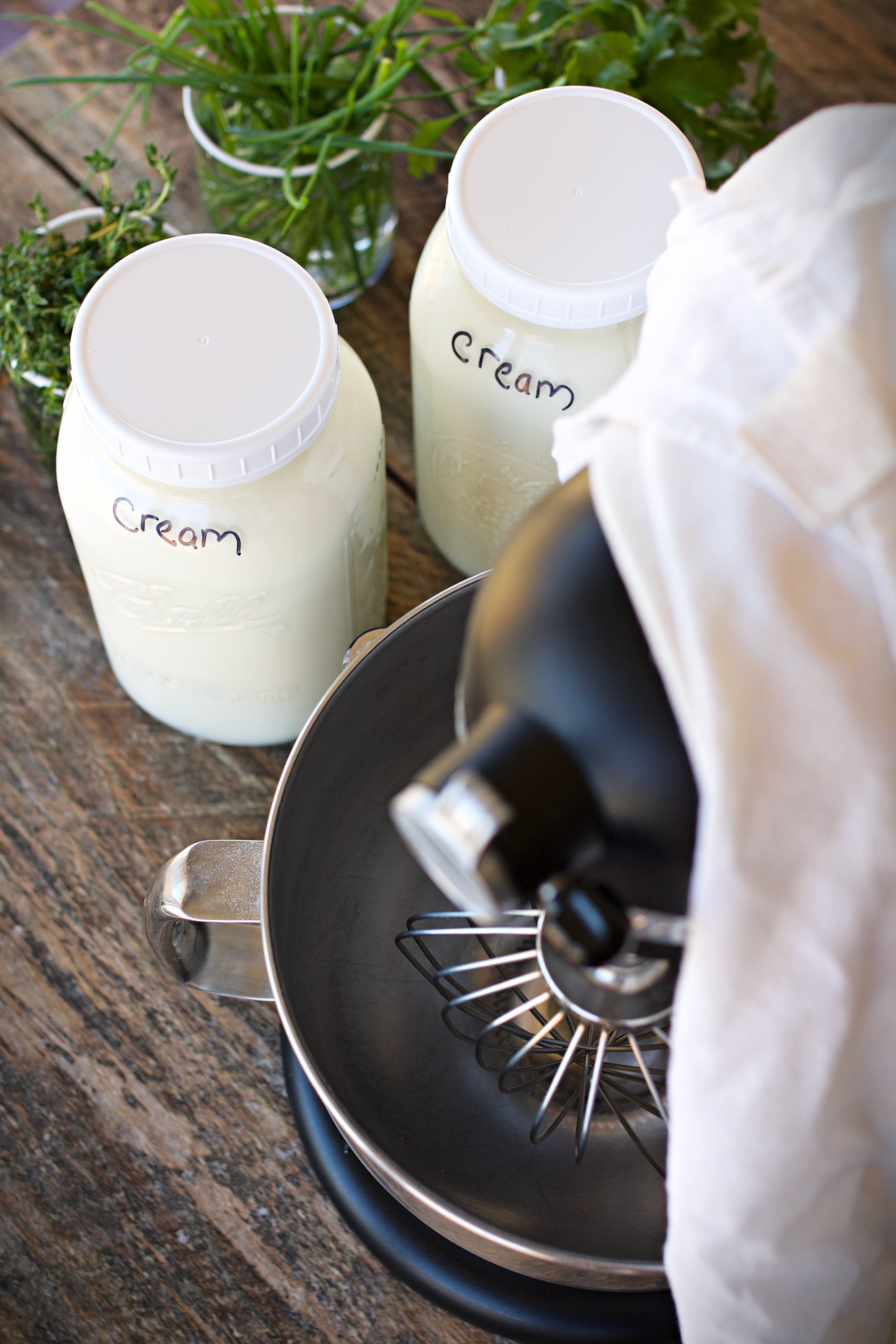 Cream in two half gallon jars next to kitchen aid that will be used to make them into butter, and herbs on the counter next to cream. 