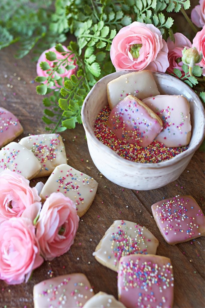 Cookies displayed in a bowl of sprinkles and on the counter with flowers.