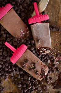 fudge popsicles laying in chocolate chips on a counter with sprinkles everywhere.