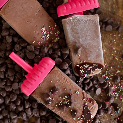 fudge popsicles laying in chocolate chips on a counter with sprinkles everywhere.