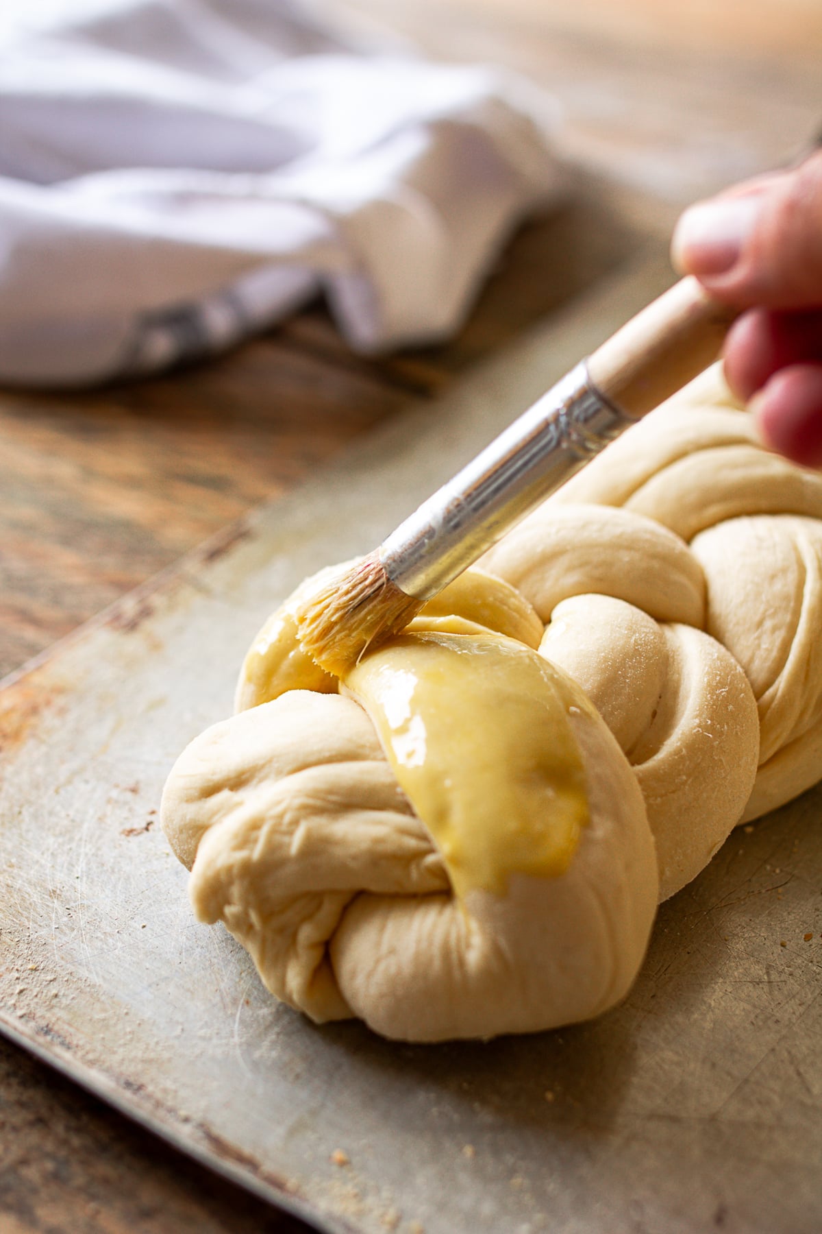 A close-up shot of a hand using a pastry brush to apply a glossy egg wash to a raw loaf of braided bread on a baking sheet.