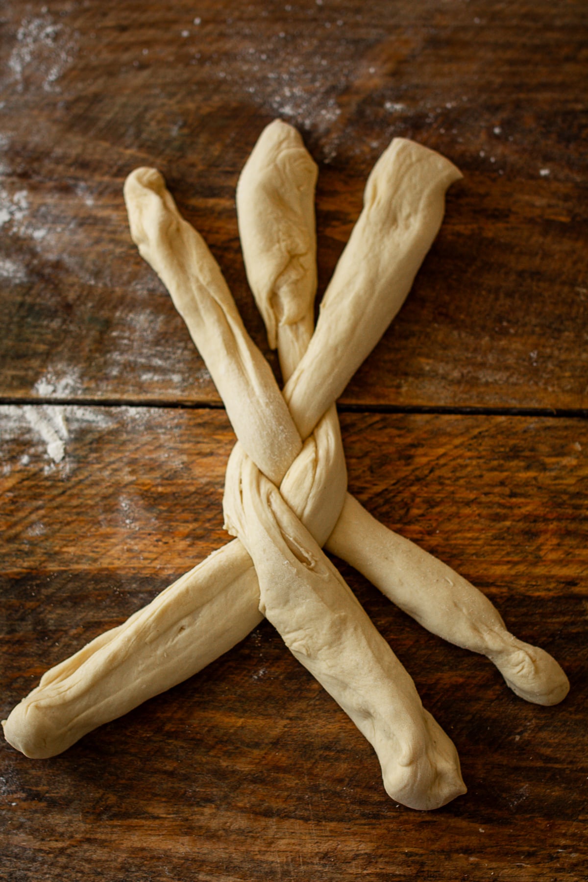 A close-up shot of three long strands of raw bread dough being braided together on a dark wooden table.
