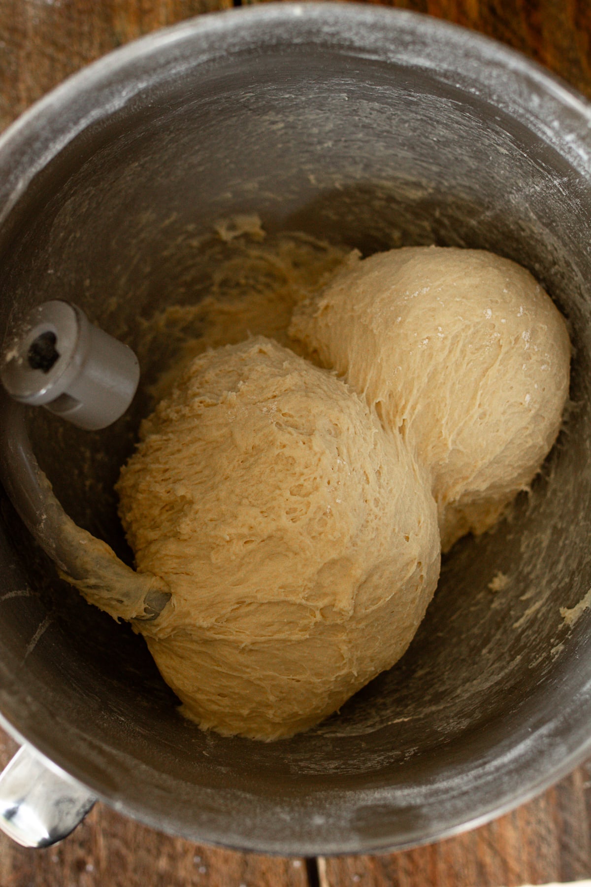 A top-down shot of a metal stand-mixer bowl containing a large ball of bread dough actively pulling away from the sides. The mixer's dough hook attachment is visible, heavily coated in dough.