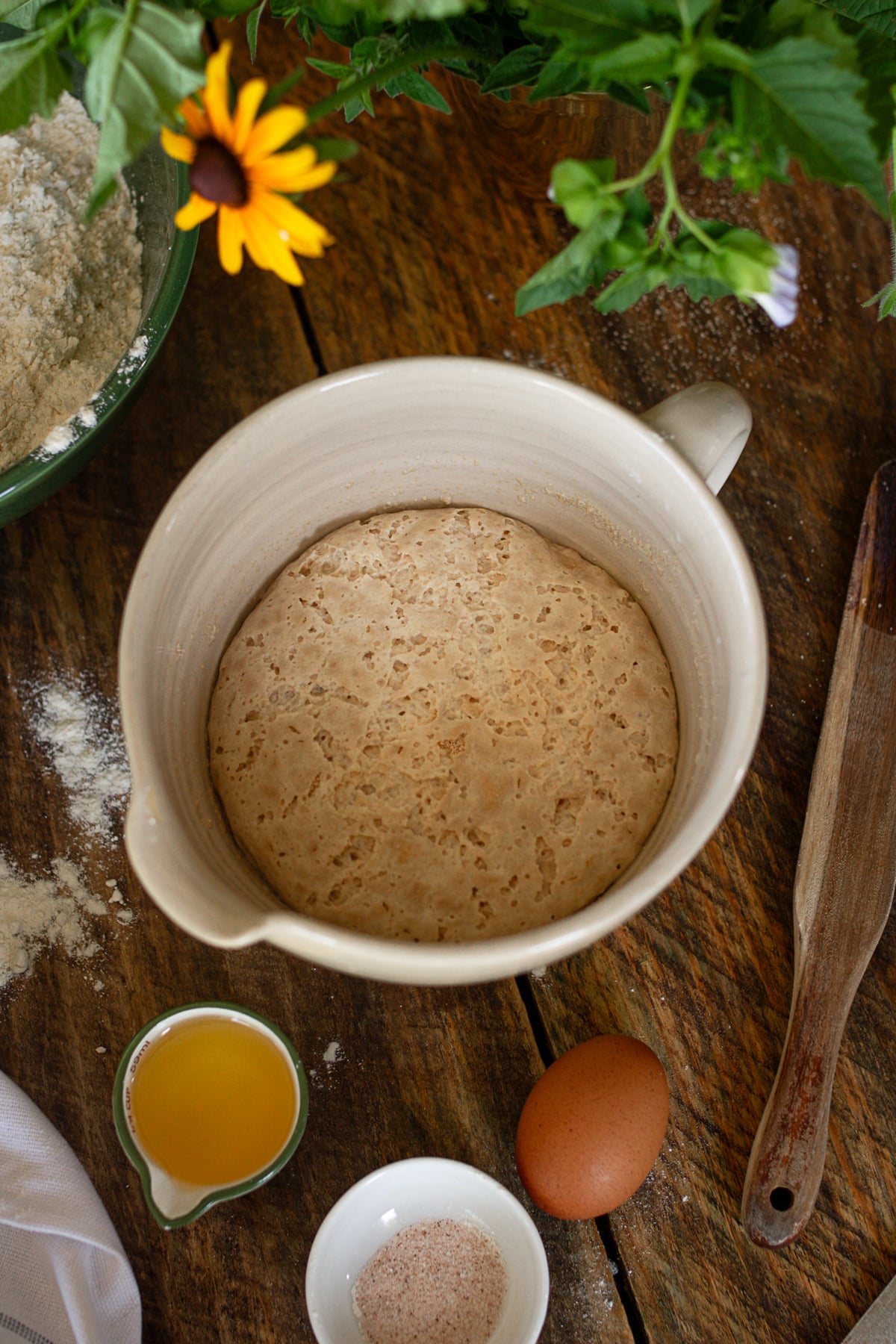 An overhead shot of bubbly yeast active in a ceramic bowl surrounded by oil, an egg, and spices on a wooden table.