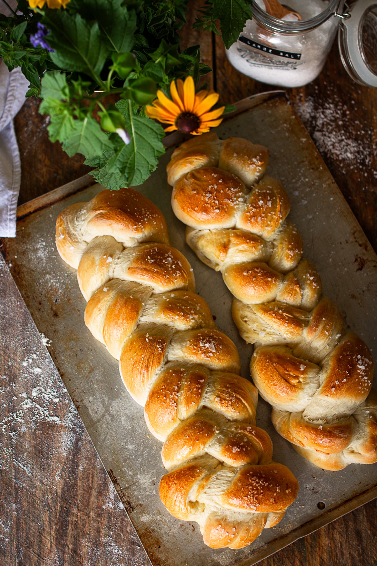 An overhead shot of two baked, golden-brown braided French bread loaves topped with coarse sea salt on a rustic metal baking sheet.