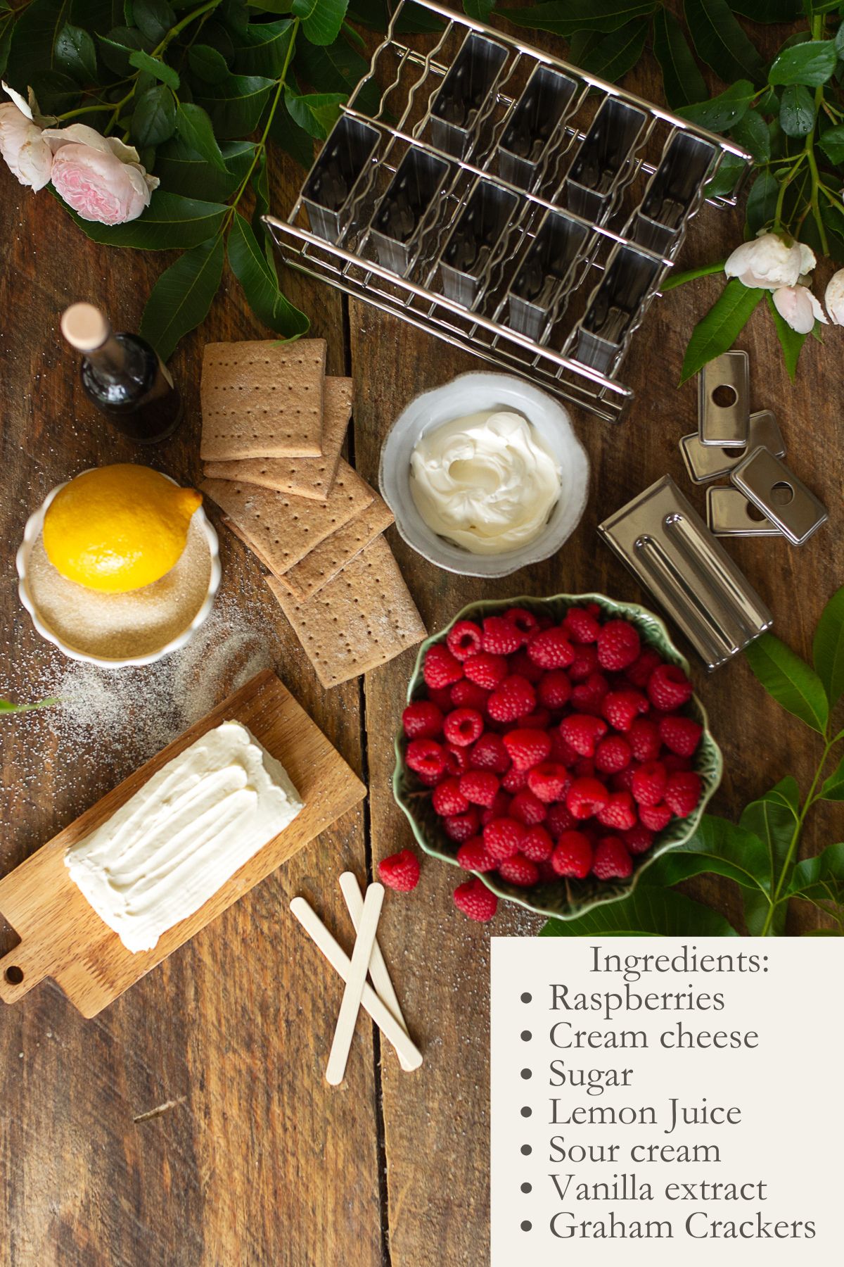 An overhead flatlay photo on a rustic wood table showing the ingredients for homemade raspberry cheesecake popsicles: a block of cream cheese, fresh red raspberries in a bowl, graham crackers, sugar, lemon, vanilla, and a stainless steel popsicle mold.