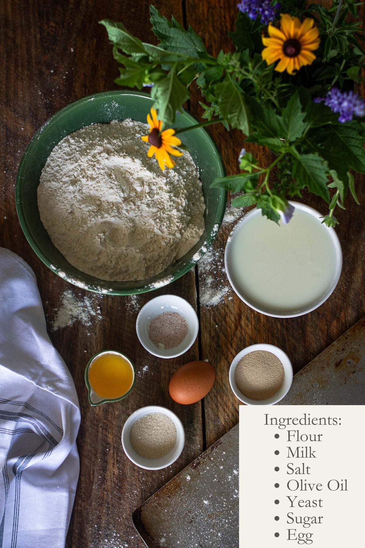 An overhead shot of baking ingredients in small bowls on a wooden table with a text box that reads: Ingredients: Flour, Milk, Salt, Olive Oil, Yeast, Sugar, Egg.