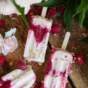A macro close-up overhead shot of two raspberry cheesecake popsicles overlapping on a wooden surface, showing the detailed red marble textures, fine graham cracker crumbs, and whole fresh berries.