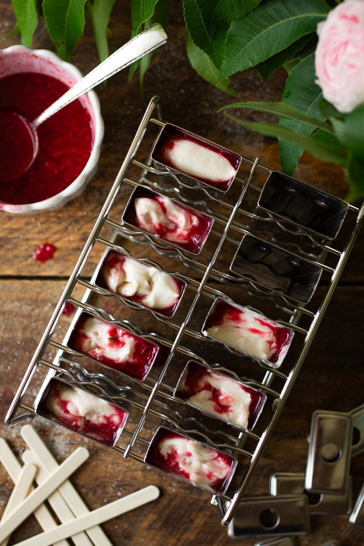 A close-up, top-down view of a stainless steel popsicle mold partially filled with layers of white cream cheese filling and vibrant red raspberry puree, showing a swirled marble effect inside the cavities.