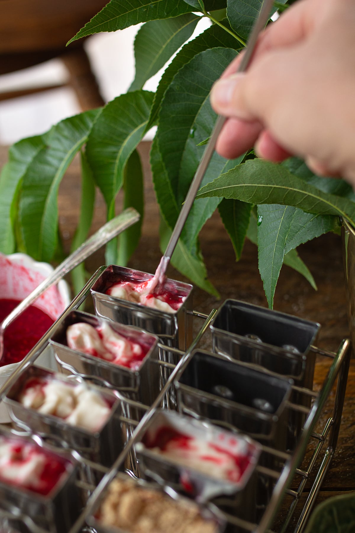 A close-up shot of a hand using a long metal skewer to gently swirl a deep red raspberry reduction into a thick, white cream cheese base inside a metal popsicle mold cavity.