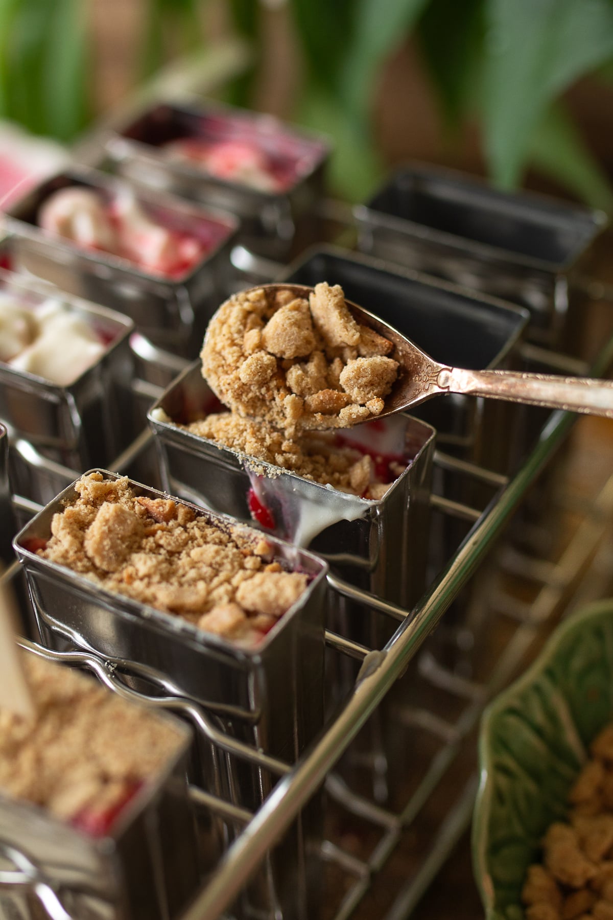 A side-angle shot of a stainless steel popsicle mold fully assembled on a wood surface, topped graham cracker crumbs, and wooden popsicle sticks.
