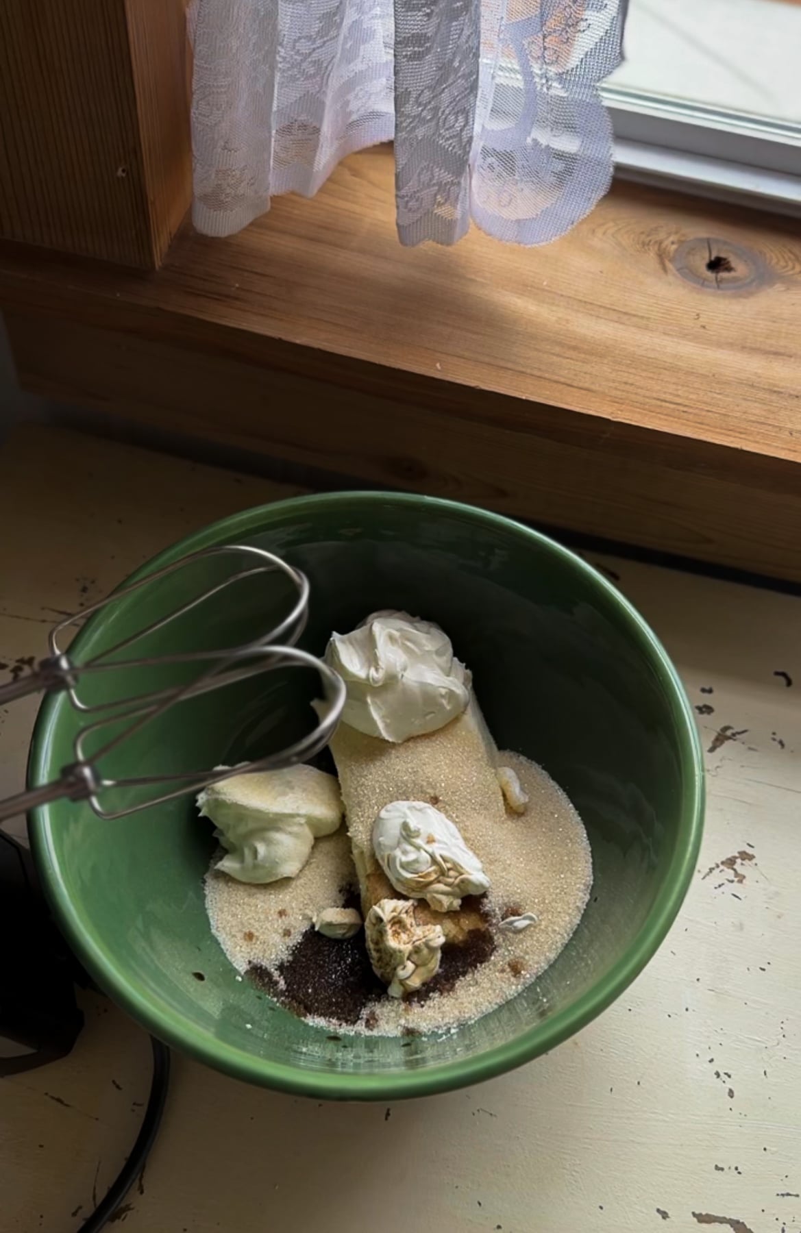 Photo looking into a green ceramic mixing bowl containing a block of cream cheese, granulated sugar, vanilla extract, and a hand mixer.