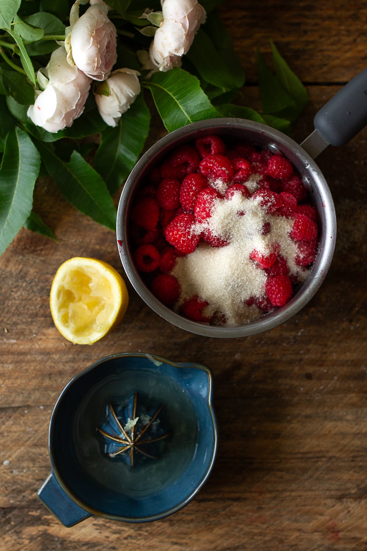 An overhead view of a small stainless steel saucepan filled with fresh red raspberries, a mound of natural cane sugar, a freshly squeezed lemon half, and a blue ceramic citrus juicer on a dark rustic wood background with leaves.