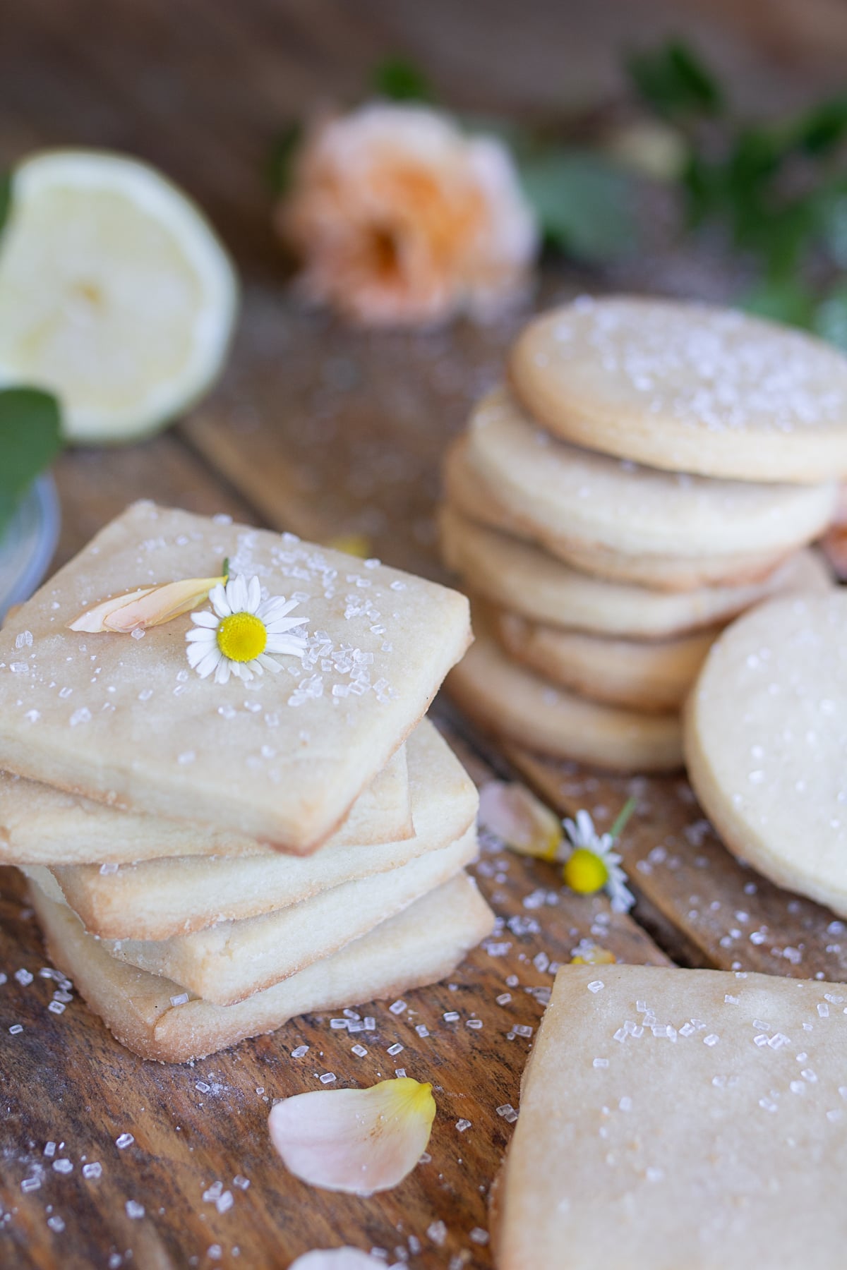 Stack of homemade square shortbread cookies topped with coarse sugar on a rustic wooden table with flowers.
