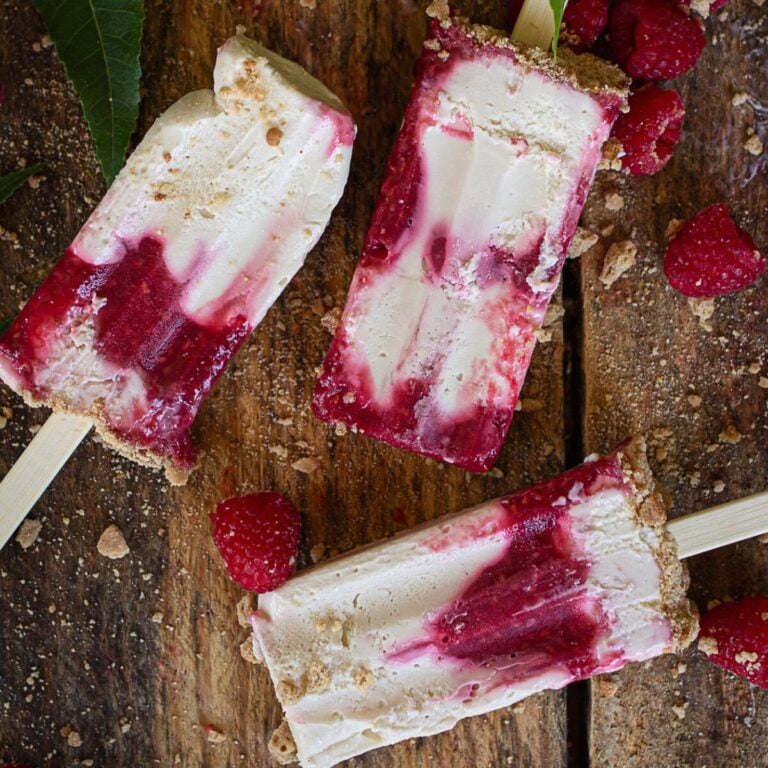 Three unmolded red and white marbled cheesecake popsicles arranged neatly on a dark wooden cutting board with fresh raspberries and graham cracker crust crumbs tucked into the corners.