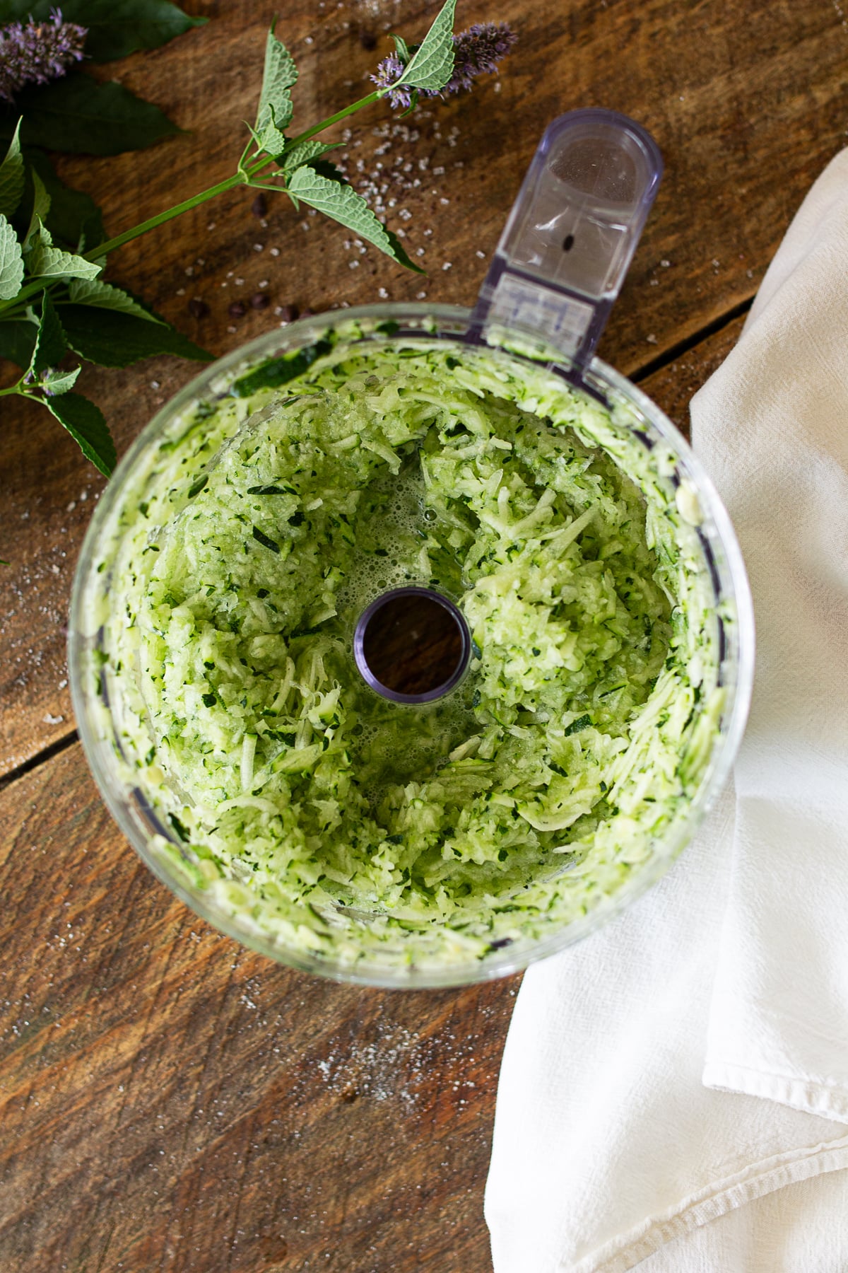Finely grated green zucchini shredded inside a clear food processor bowl on a wooden countertop.