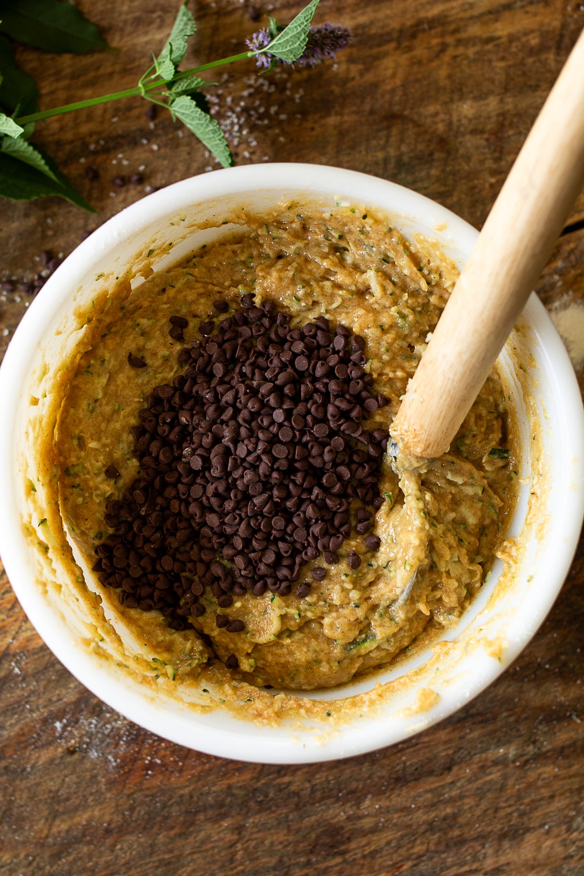 Thick spiced zucchini bread batter in a white mixing bowl topped with a cup of mini chocolate chips and a wooden spoon.