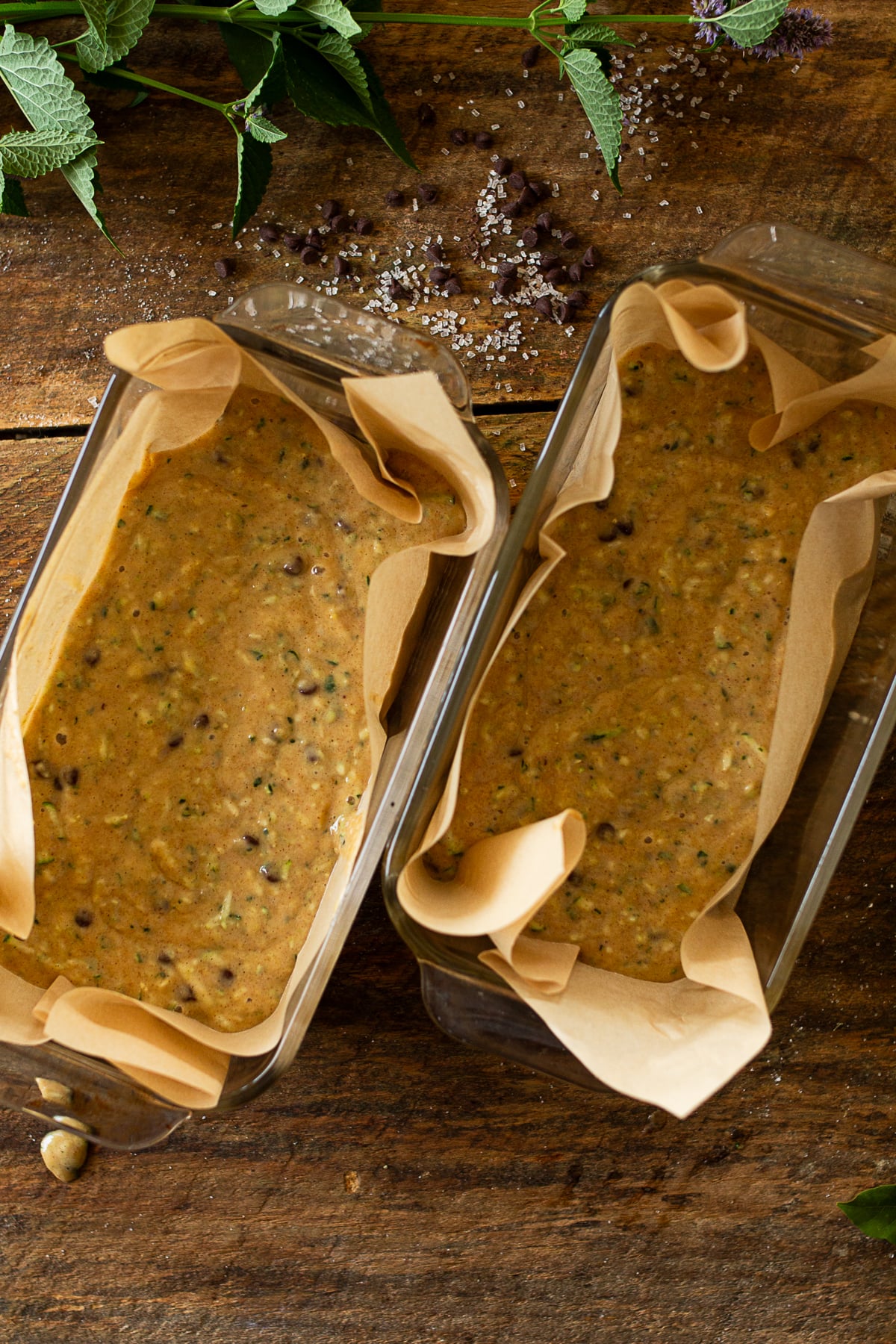Two parchment lined glass loaf pans filled with raw chocolate chip zucchini bread batter on a rustic wood background.