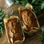 Two freshly baked golden brown loaves of farmhouse chocolate chip zucchini bread cooling in parchment lined glass pans on a rustic wooden table.