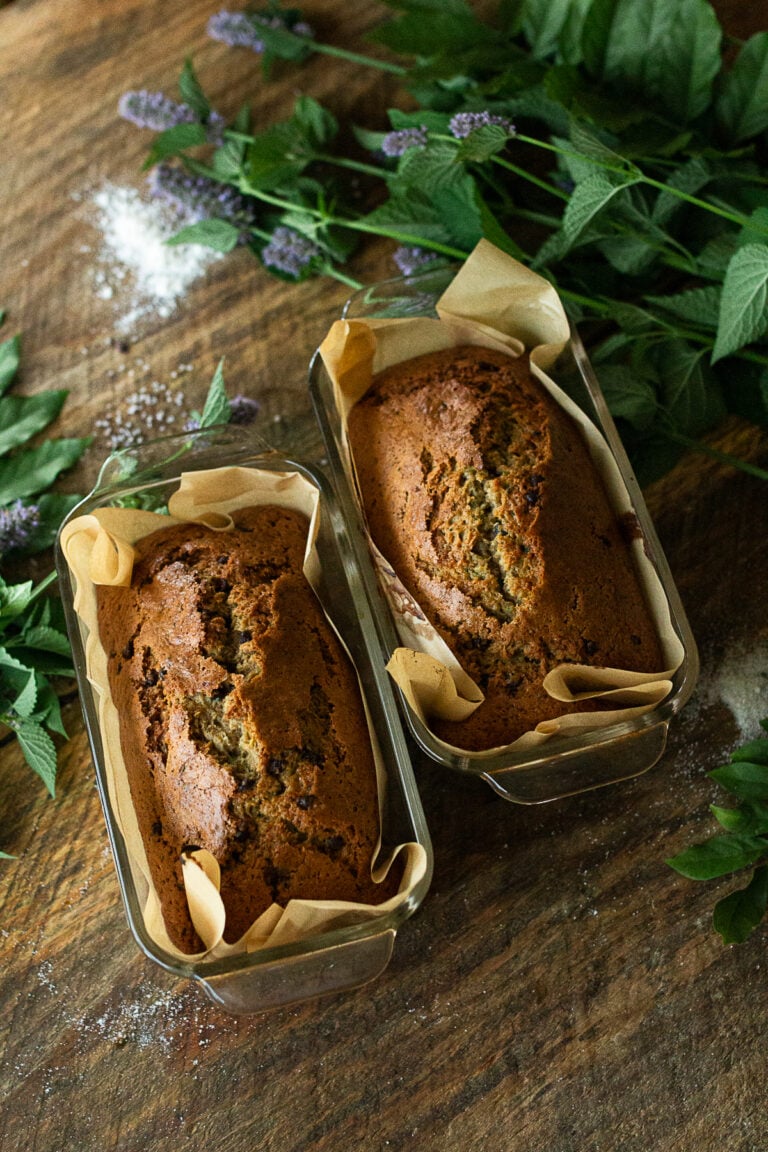 Two freshly baked golden brown loaves of farmhouse chocolate chip zucchini bread cooling in parchment lined glass pans on a rustic wooden table.