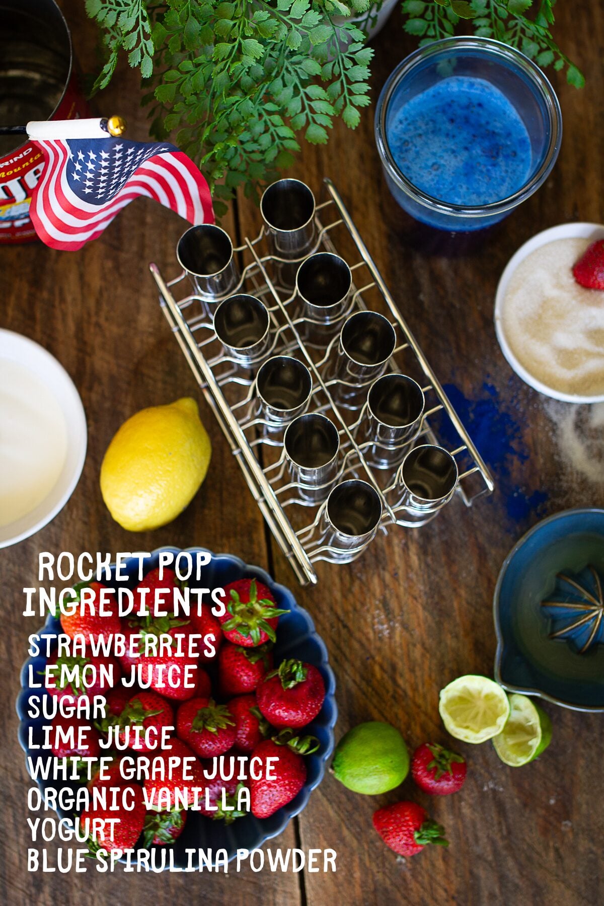 An overhead photograph on a rustic wood table showing ingredients for homemade rocket pops, including a blue bowl of strawberries, whole lemons and limes, yogurt, white grape juice in a jar, blue spirulina powder, a citrus juicer, and a 10-cavity stainless steel popsicle mold. Text overlay lists "ROCKET POP INGREDIENTS."