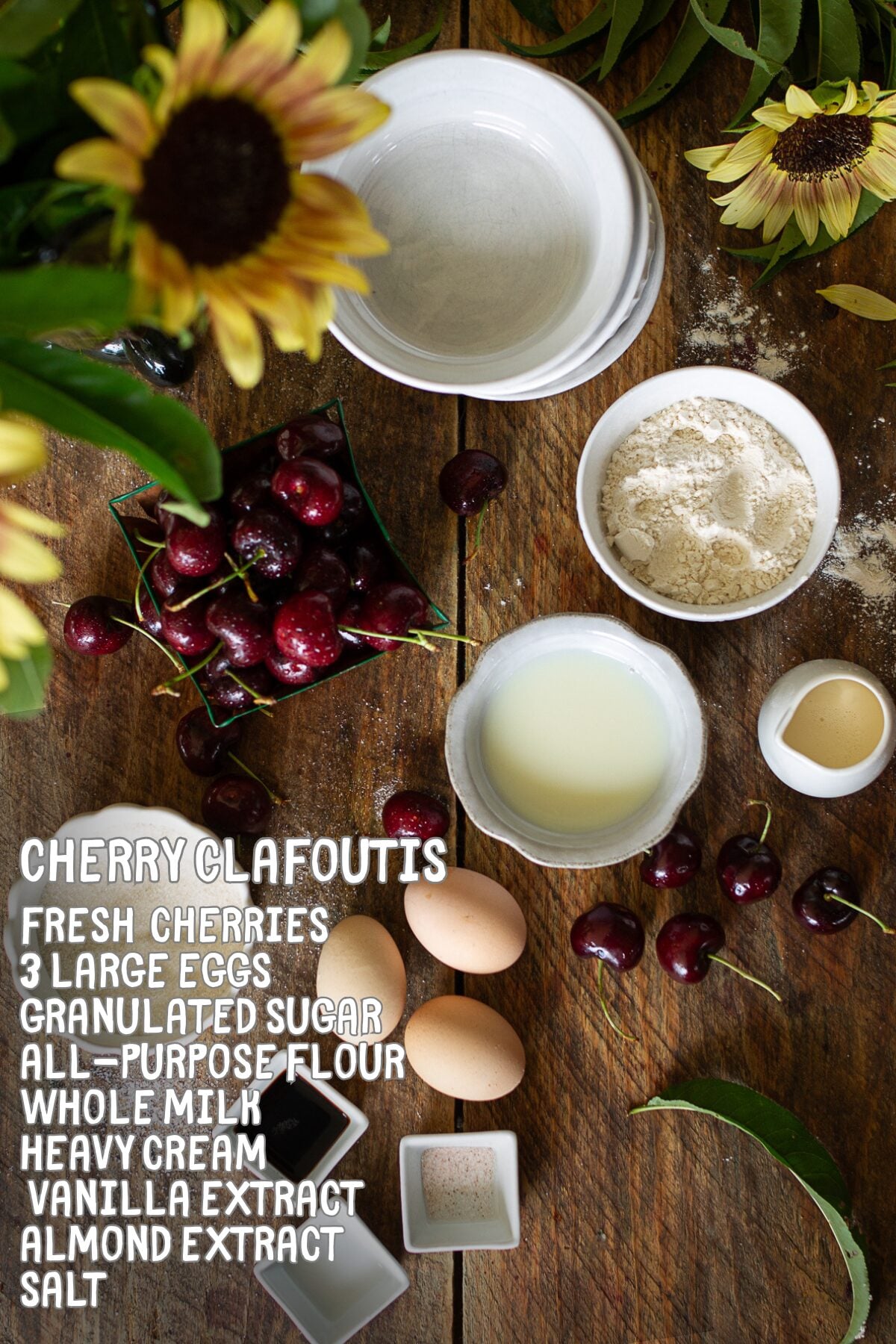 Flat lay ingredients for traditional French cherry clafoutis recipe including fresh sweet cherries, farm eggs, whole milk, heavy cream, flour, and sugar on wood background.