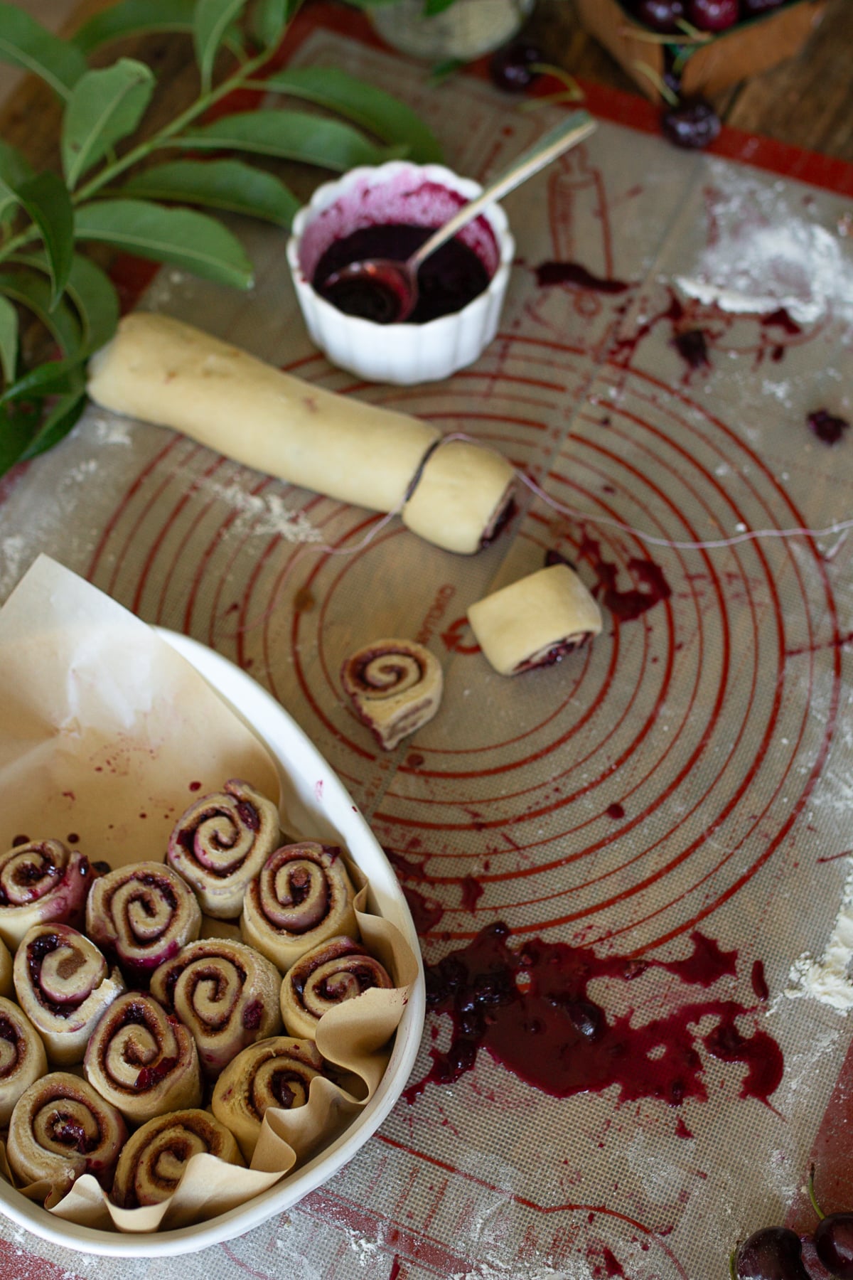 Sourdough cinnamon roll dough log being sliced with string alongside unbaked rolls lined in a parchment baking dish