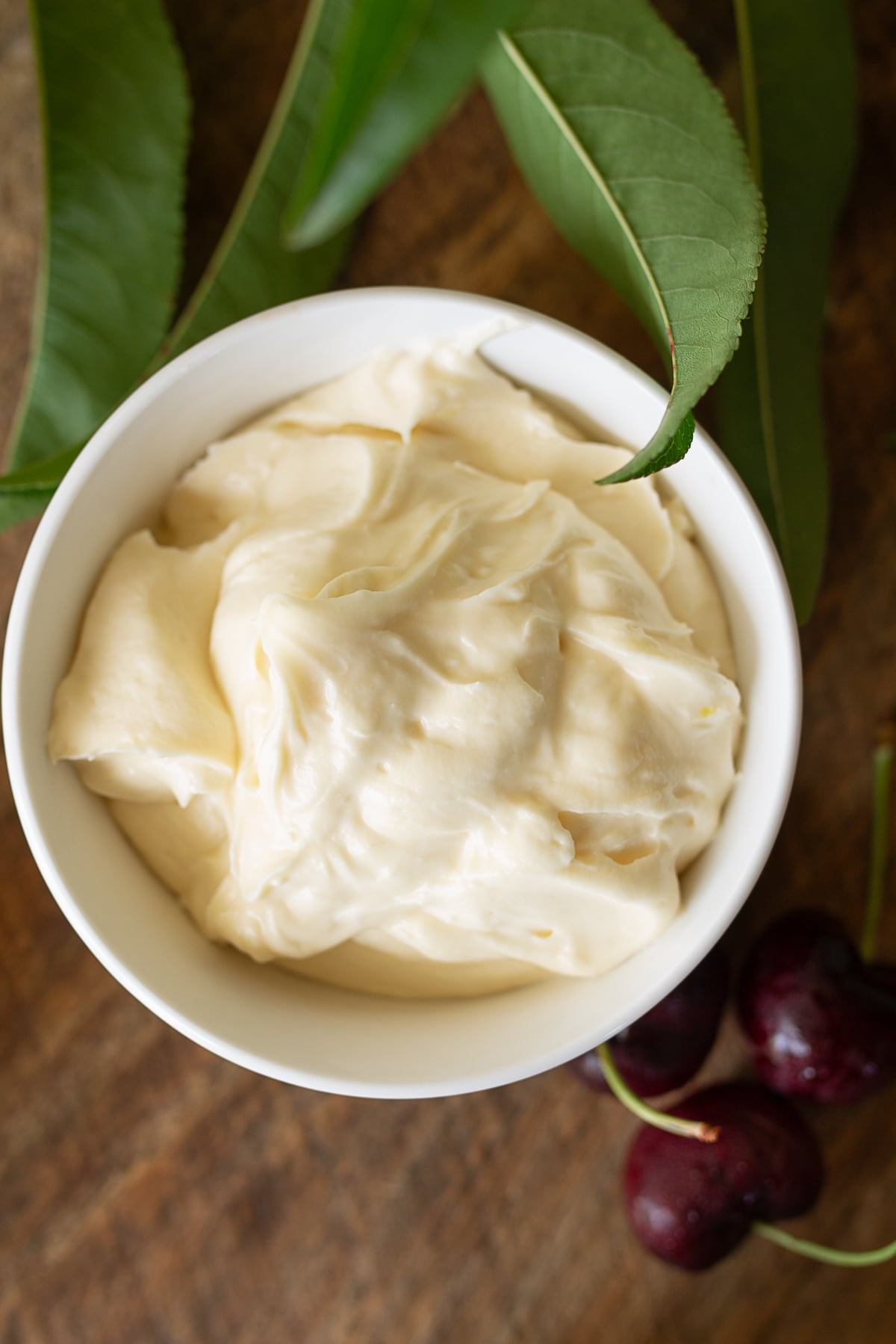 Overhead view of smooth whipped cream cheese frosting in a small ceramic bowl next to fresh cherries