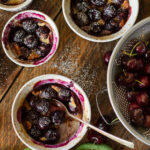 Top down view of individual cherry clafoutis in white ramekins on a rustic wooden table with fresh sweet cherries in a colander.