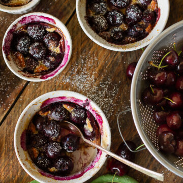 Top down view of individual cherry clafoutis in white ramekins on a rustic wooden table with fresh sweet cherries in a colander.