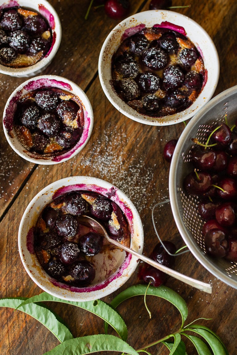 Top down view of individual cherry clafoutis in white ramekins on a rustic wooden table with fresh sweet cherries in a colander.