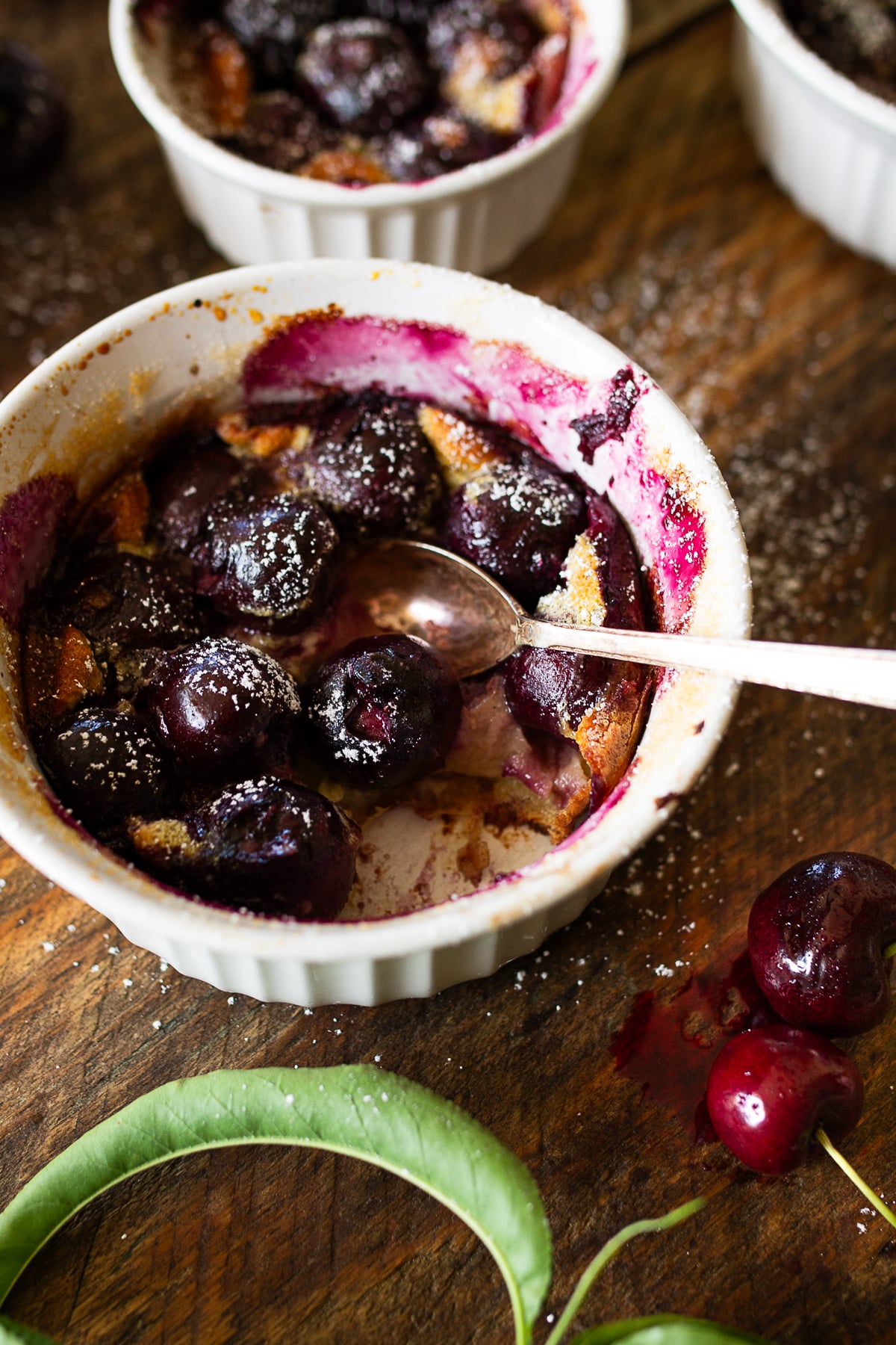 Close up of a single serving individual cherry clafoutis with a silver spoon digging into the golden custard cake next to fresh dark sweet cherries.