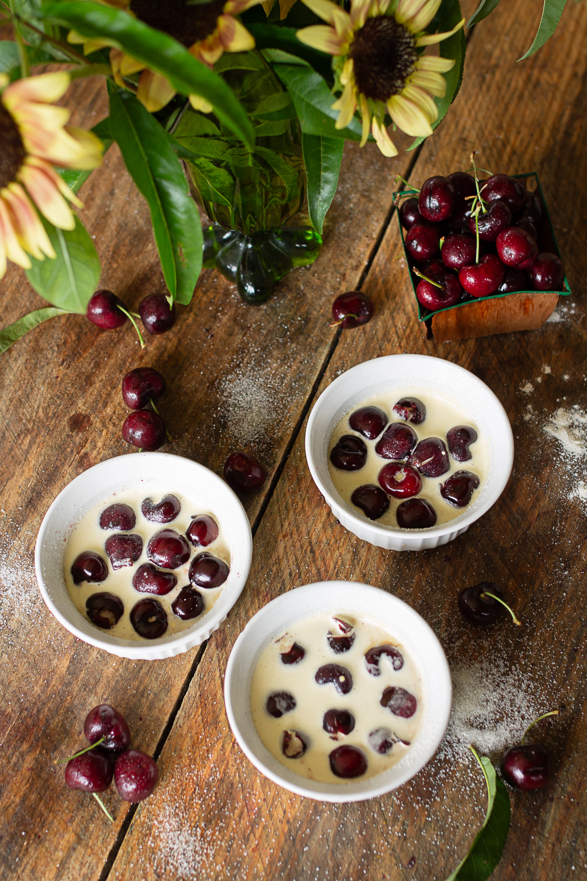 Three white ramekins filled with sweet cherries and raw liquid custard batter ready to bake on a farmhouse wooden table next to fresh sunflowers.