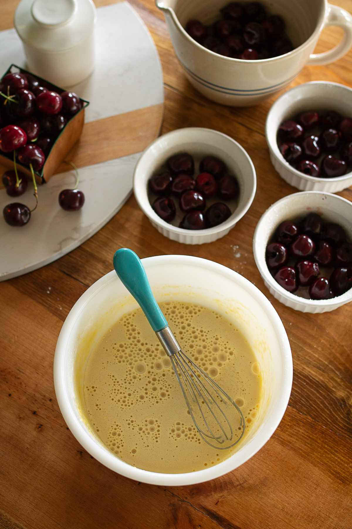 Smooth golden custard batter in a white mixing bowl with a teal whisk next to ramekins filled with fresh pitted sweet cherries.
