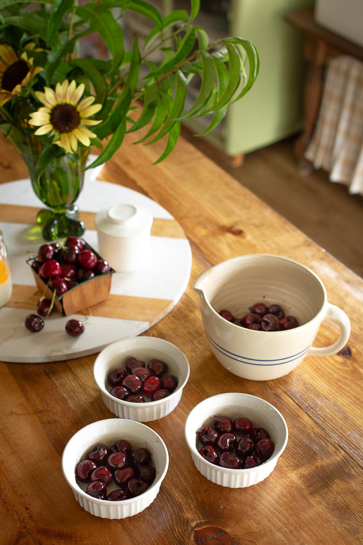 Cherries in small bowls next to large bowl of cherries on kitchen counter with sunflowers in background.